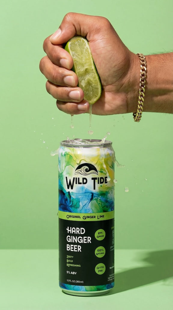 A hand with a gold chain squeezes a fresh lime over a can of Wild Tide Original alcoholic ginger beer with a green background.