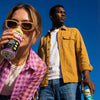 Two people holding Wild Tide hard ginger beer cans against a clear blue sky.