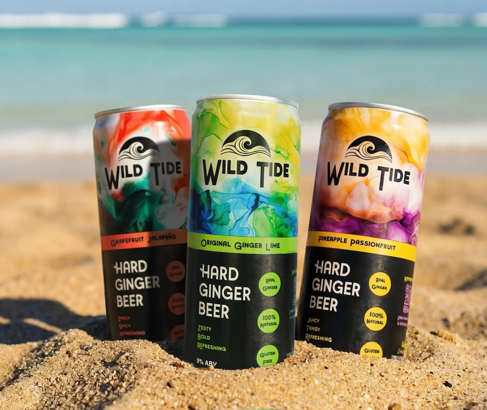 Three cans of Wild Tide Hard Ginger Beer on a sandy beach with ocean in the background