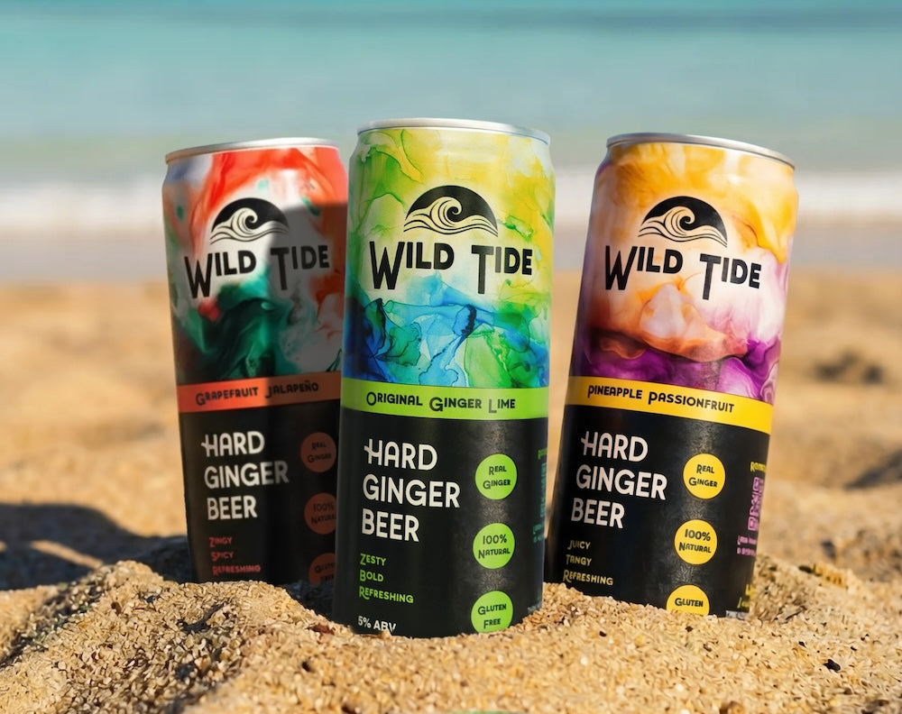 Three cans of Wild Tide Hard Ginger Beer on a sandy beach with ocean in the background
