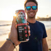A guy with a bracelet on the beach holds out a can of Wild Tide Grapefruit Jalapeño hard ginger beer with a guy wearing a blue shirt smiling in the background.