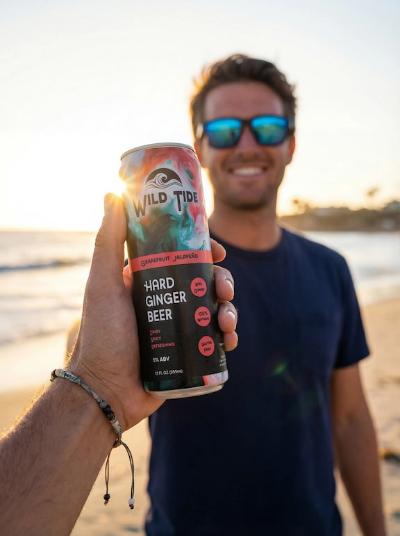 A guy with a bracelet on the beach holds out a can of Wild Tide Grapefruit Jalapeño hard ginger beer with a guy wearing a blue shirt smiling in the background.