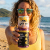 Woman with glasses stands at the beach in a yellow top holding a can of Wild Tide Pineapple Passionfruit toward the camera.