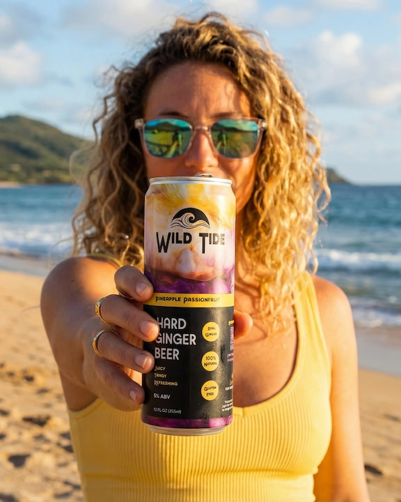 Woman with glasses stands at the beach in a yellow top holding a can of Wild Tide Pineapple Passionfruit toward the camera.