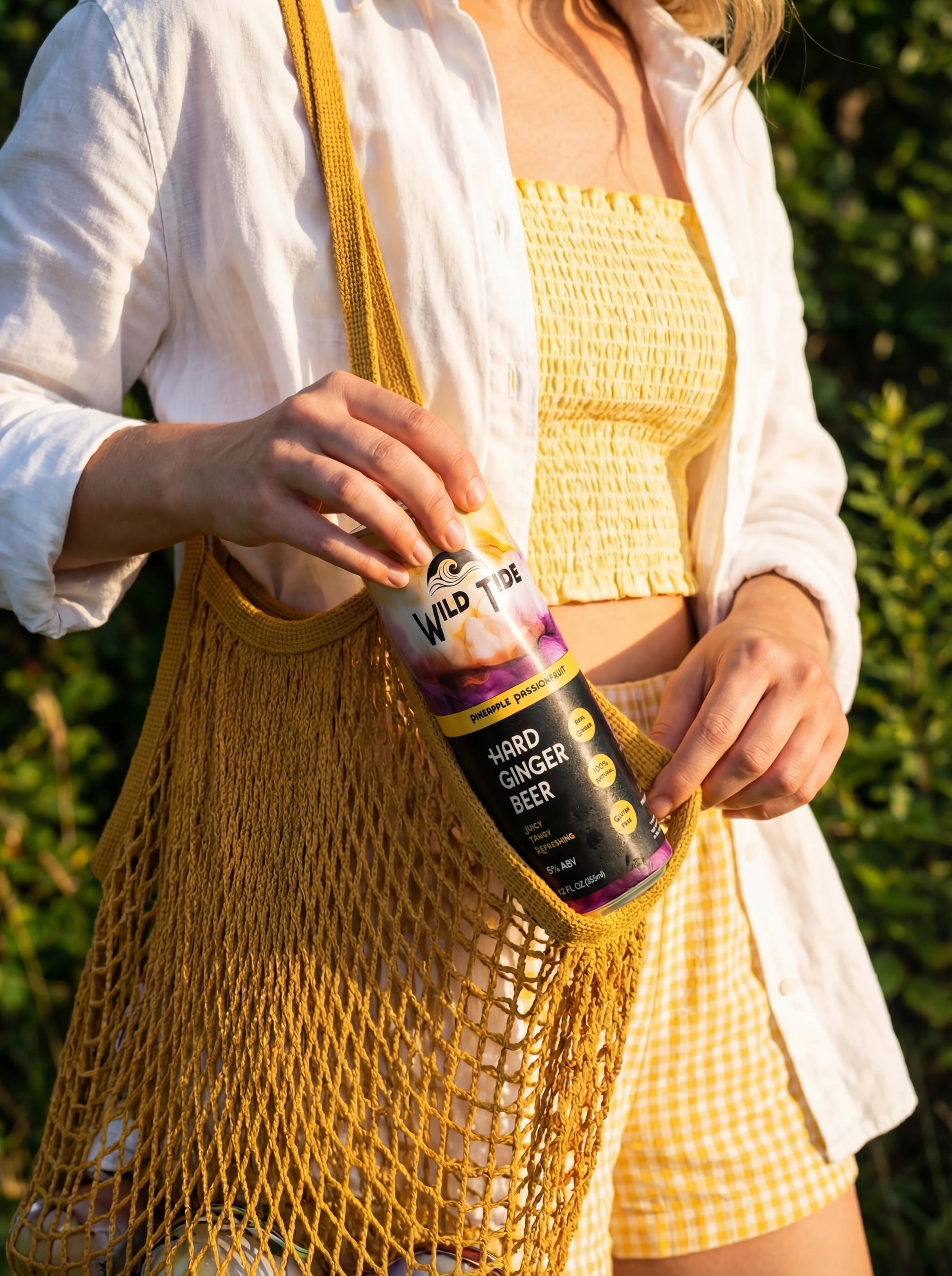 Woman in a yellow shirt and shorts and a white cover top holds a yellow sling bag pulling out a can of Wild Tide Pineapple Passionfruit ginger beer.