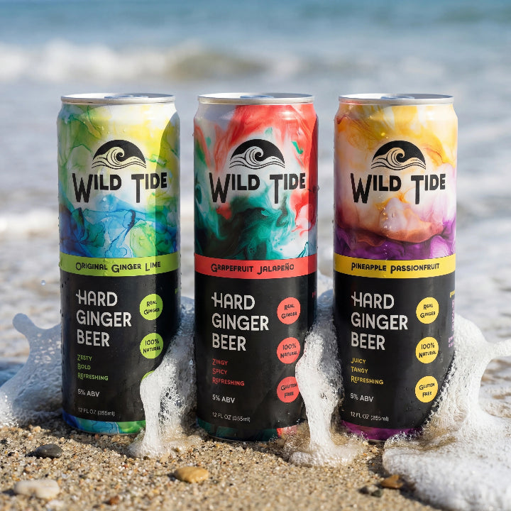 Three cans of Wild Tide hard ginger beer on the beach with a wave hitting them from the back.
