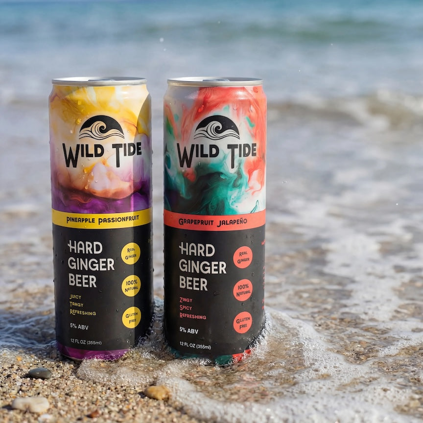 Two cans of Wild Tide Hard Ginger Beer on a beach with ocean waves in the background.